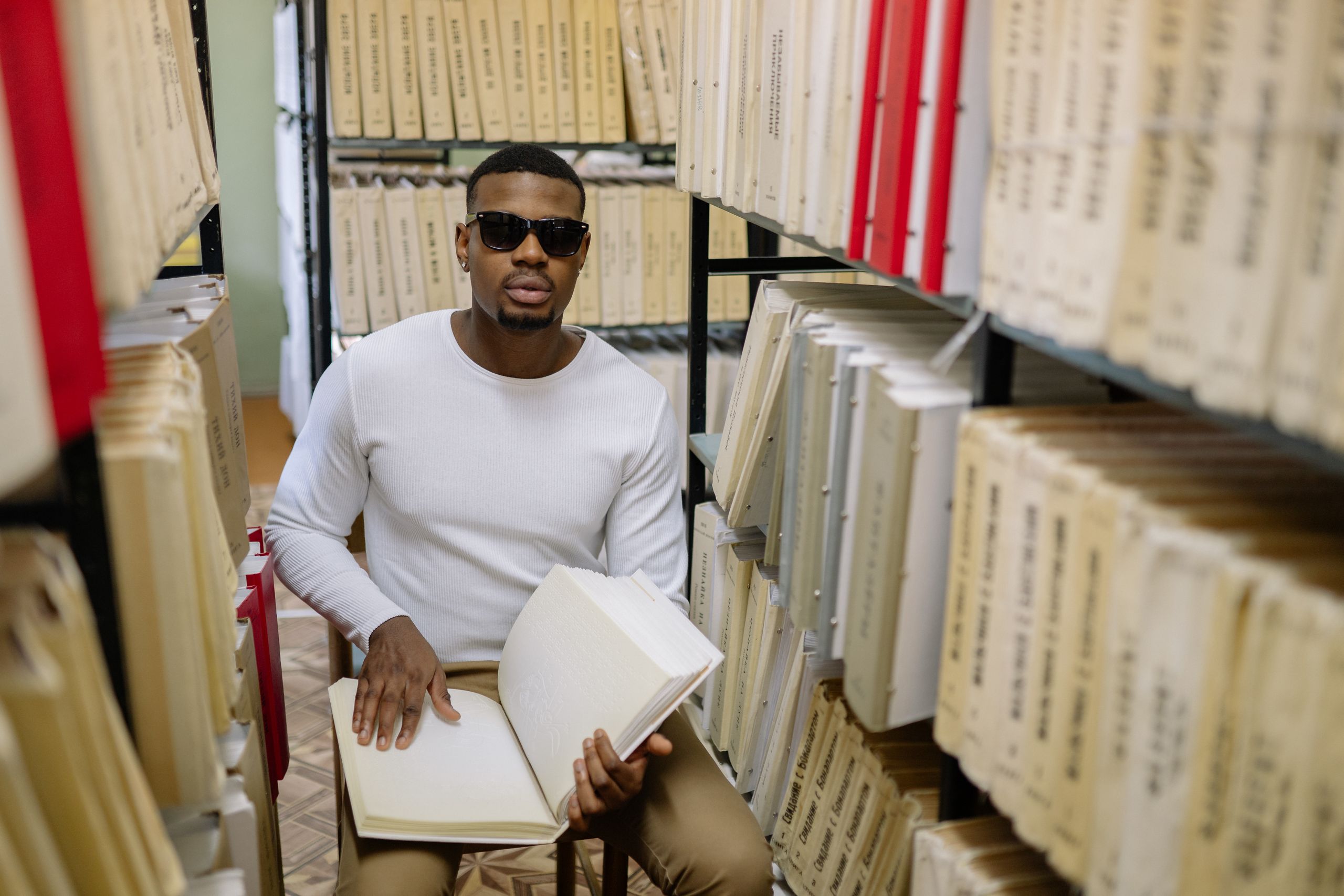A blind man reading a braille book in a quiet library setting, surrounded by shelves.